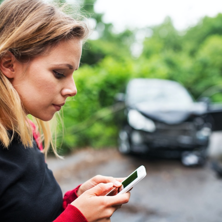 A young woman with smartphone by the damaged car after a car accident, text messaging.