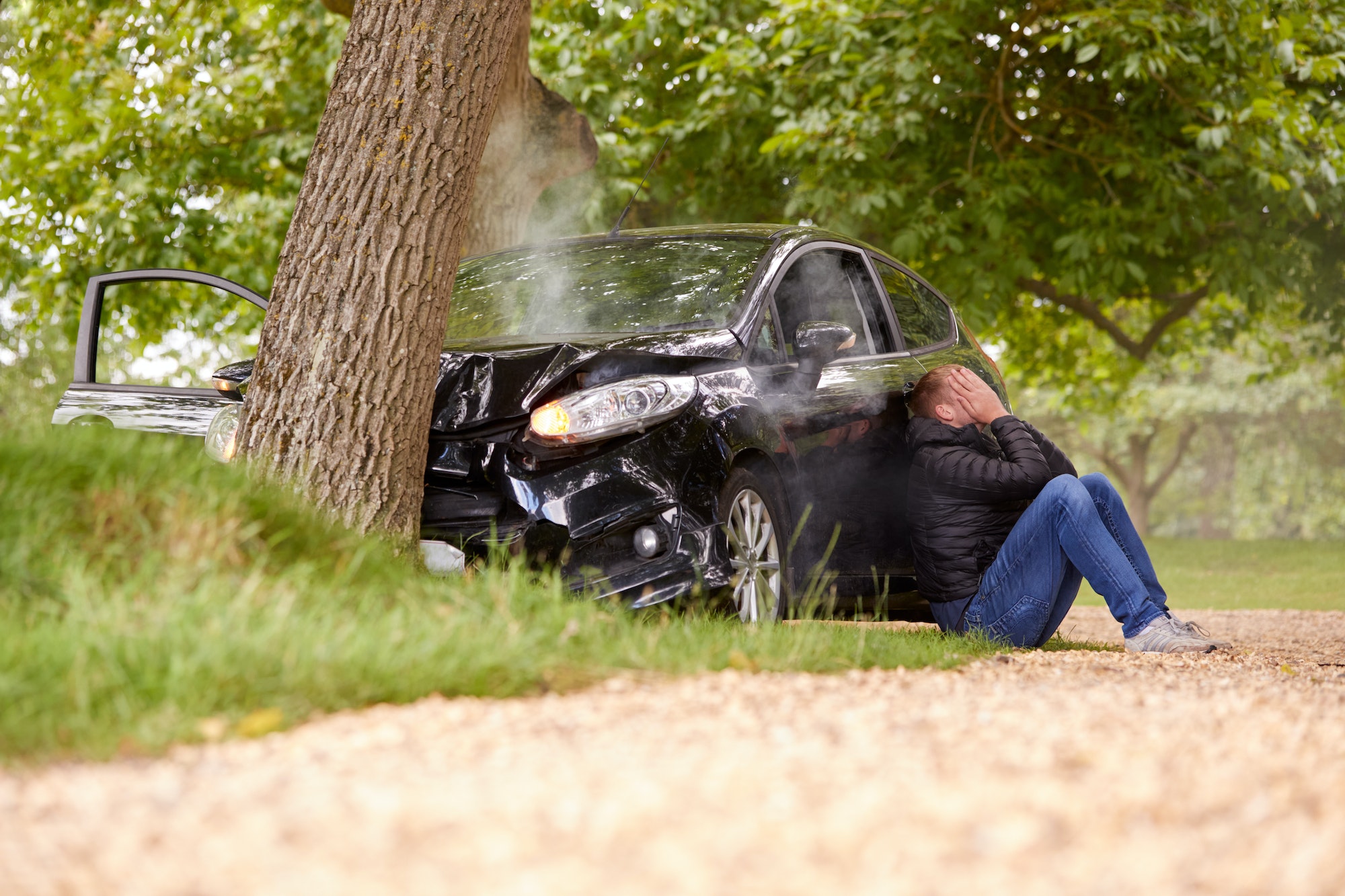 Teenage Driver With Head In Hands Sitting Next To Wrecked Car After Crashing Into Tree